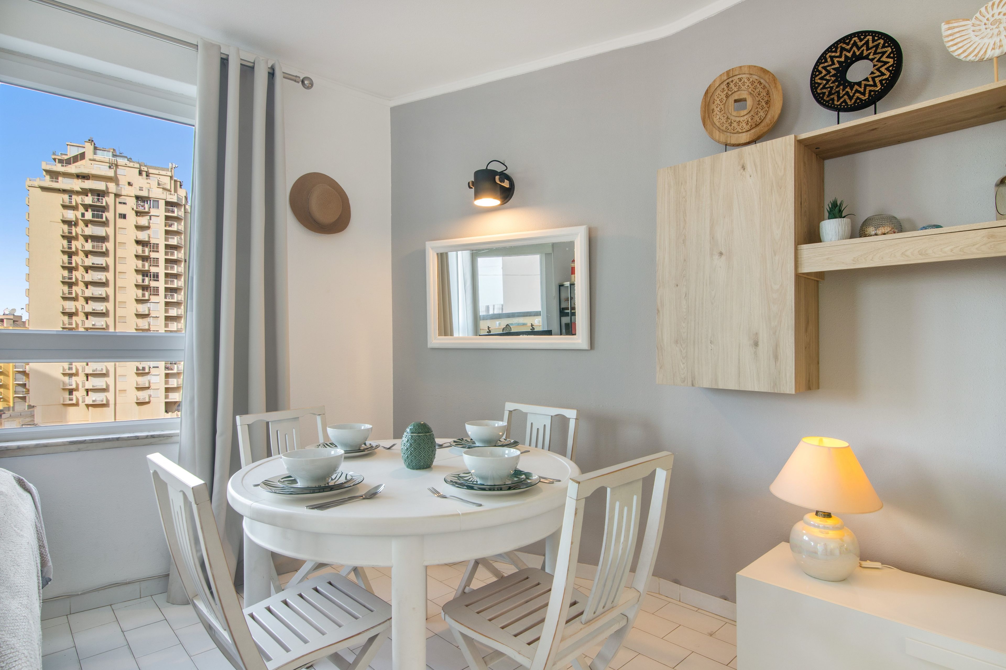 A dining area with a round table, white chairs, and a view of a high-rise building through a window.