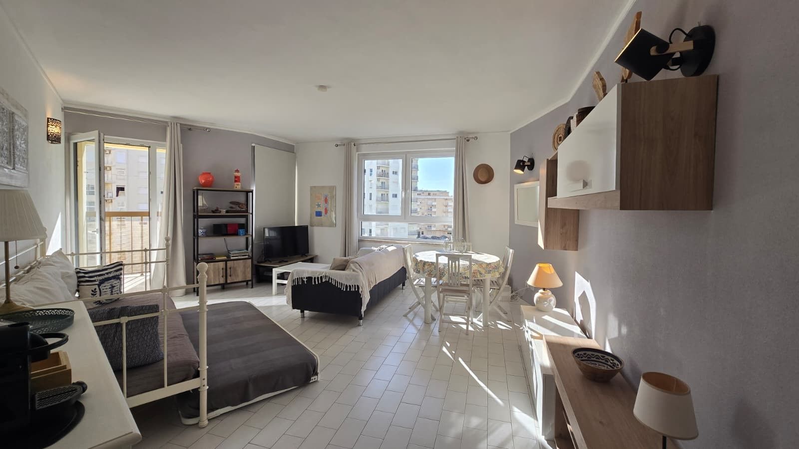 Ein helles Zimmer mit einem Bett, einem Schreibtisch, einem Sessel, einem Fernseher und einem Esstisch mit Stühlen.