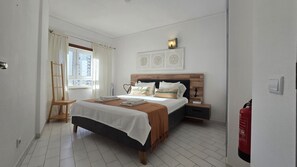 Ein Hotelzimmer mit Bett, Nachttischen, einem Fenster mit Vorhängen, einer Wandlampe und einem Feuerlöscher.