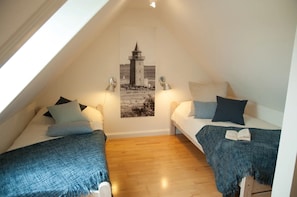 Une petite chambre mansardée, chaleureuse et accueillante, avec deux lits simples, un plancher en bois et une photographie de phare accrochée au mur.