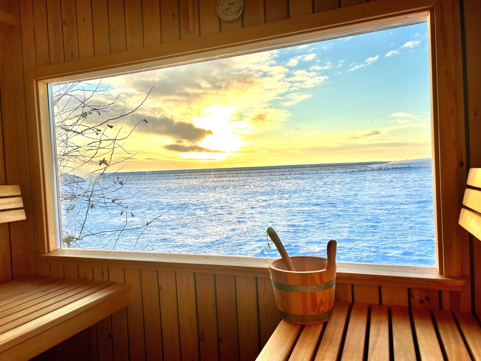 Une cabane en bois dotée d’une grande fenêtre donnant sur une étendue d’eau au coucher du soleil.