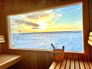 Une cabane en bois dotée d’une grande fenêtre donnant sur une étendue d’eau au coucher du soleil.