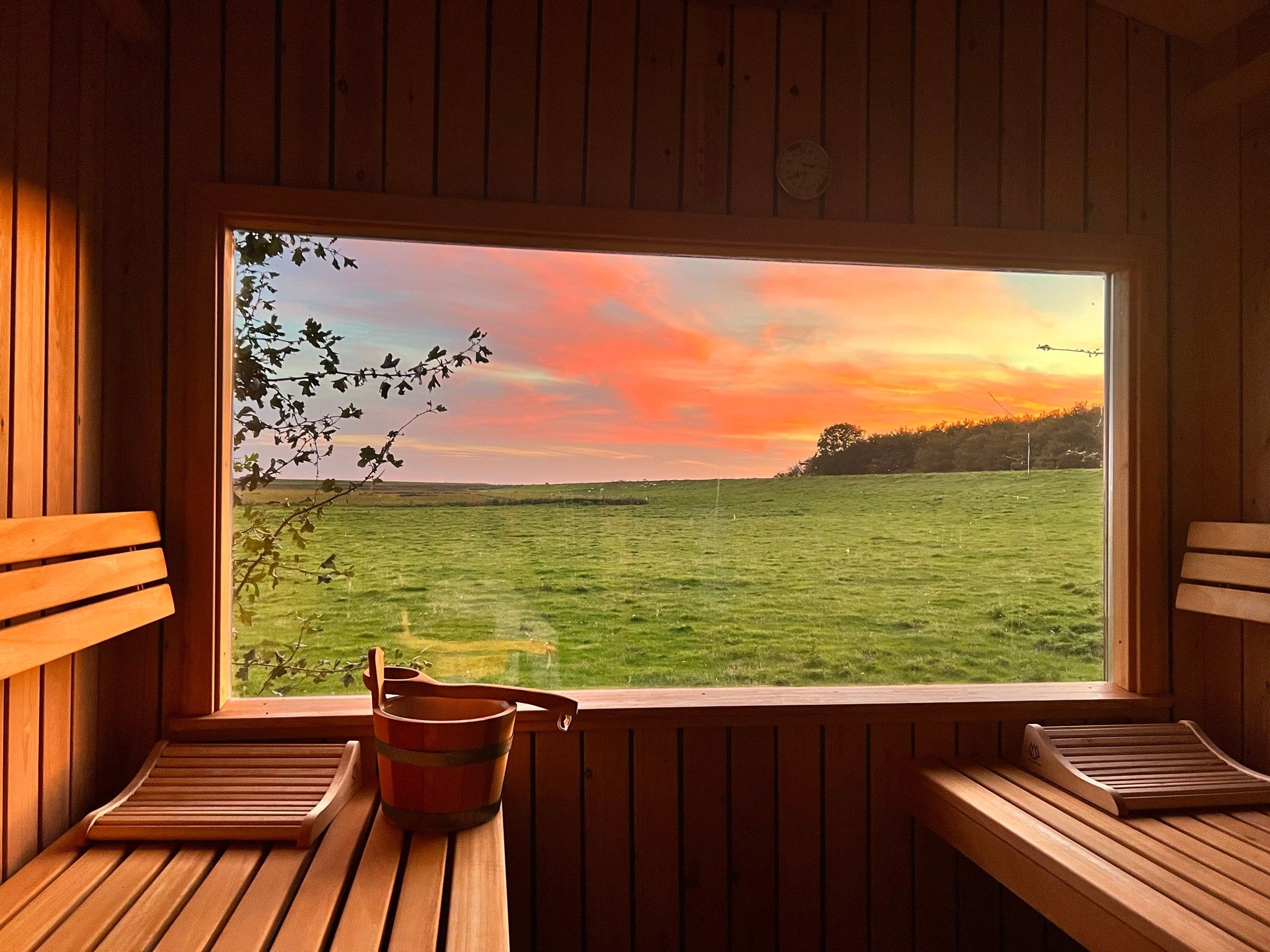 Eine Holzhütte mit einem großen Fenster mit Blick auf ein saftiges Grasfeld und einen Sonnenuntergang.