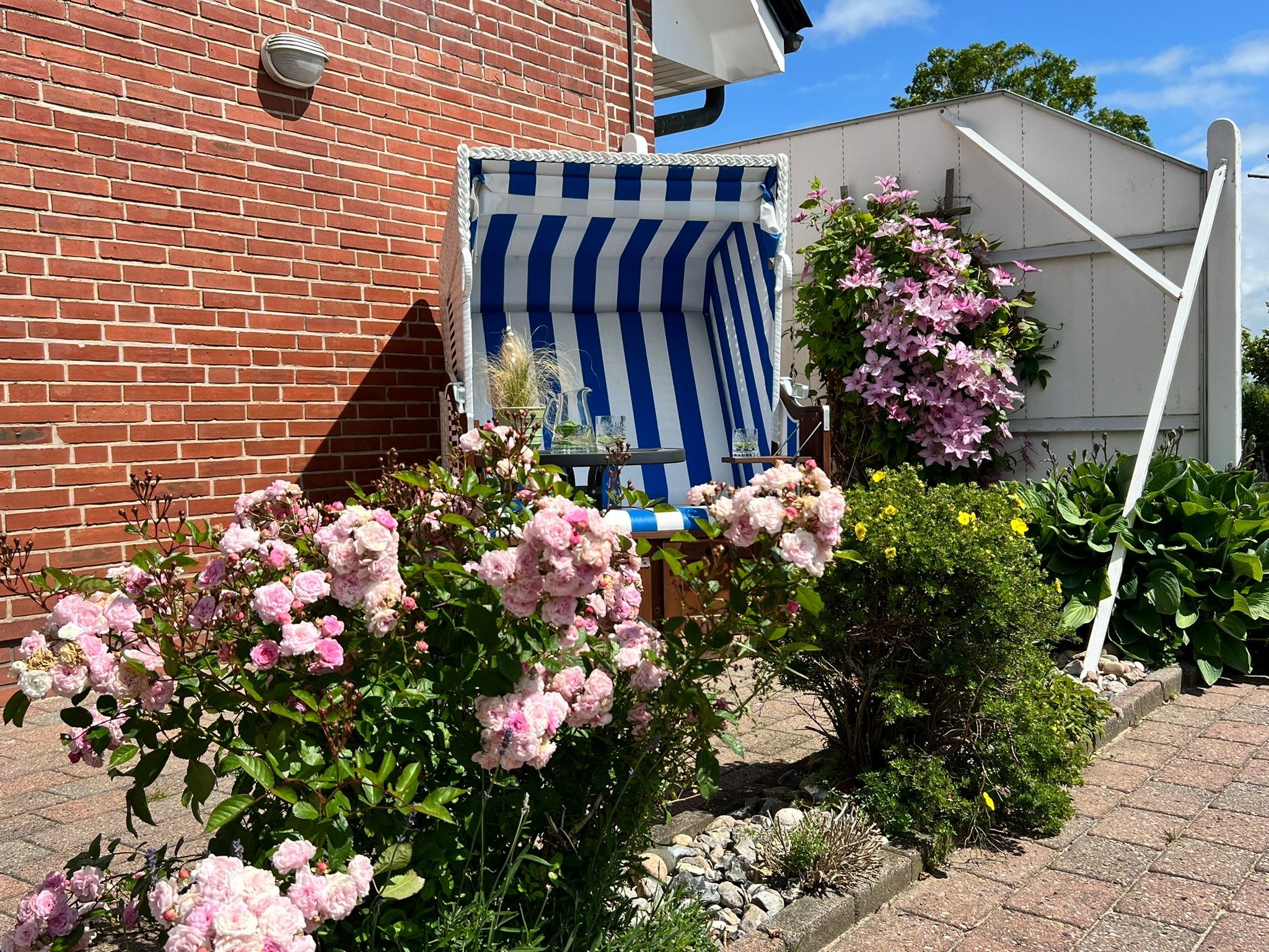 Un bâtiment en briques, avec une chaise à rayures bleues et blanches devant, entouré de fleurs roses et de verdure.