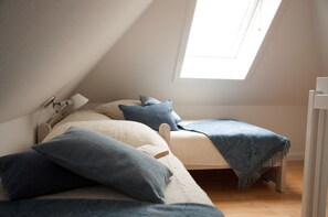 Une petite pièce agréable, avec un plafond en pente et un puits de lumière. On y trouve deux lits avec des oreillers et une couverture, une table de chevet avec une lampe, et un plancher en bois.