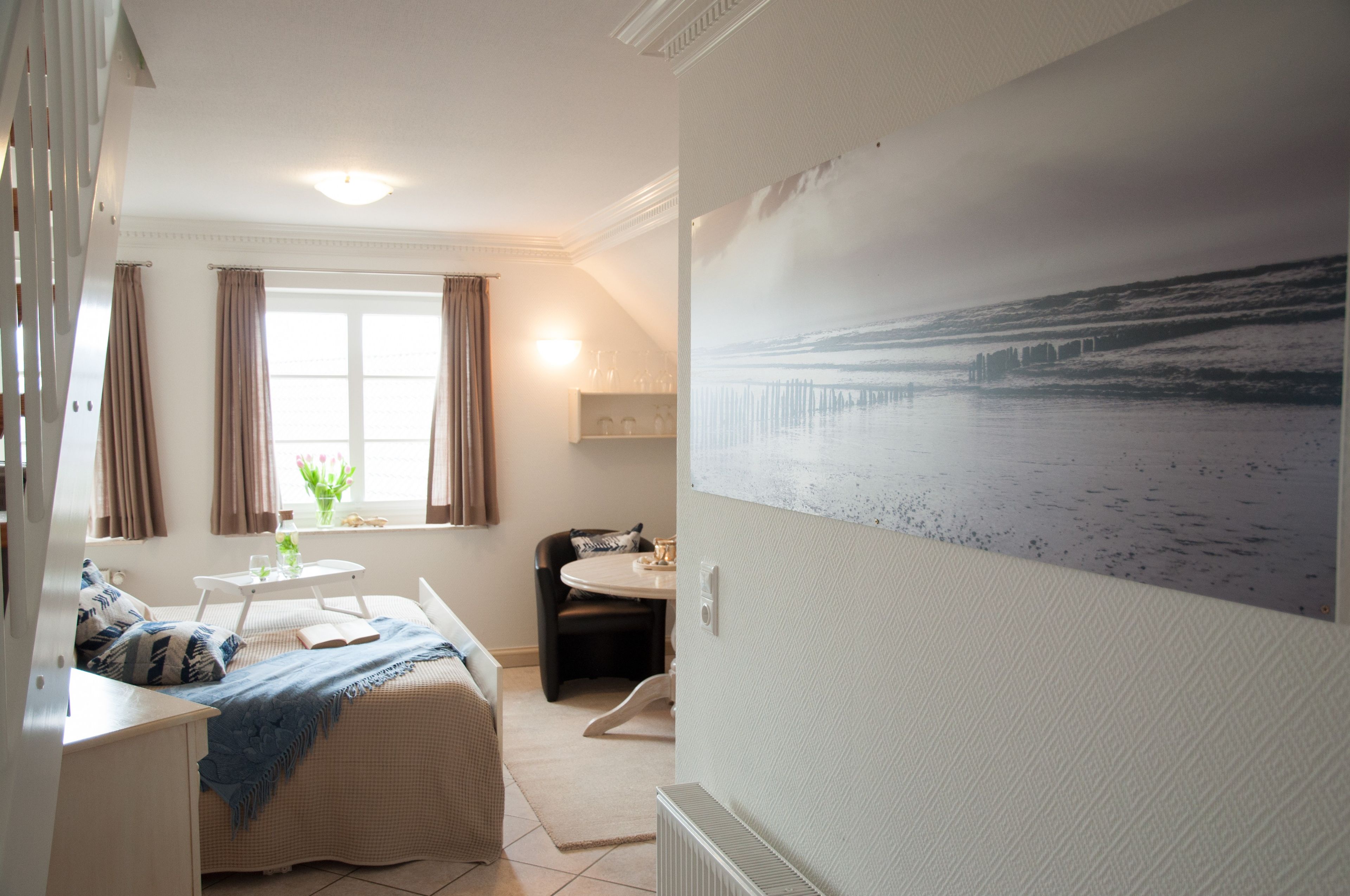 A room with a bed, a small table, a chair, a window with curtains, and a wall-mounted painting of a beach scene.