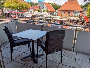 Ein Balkon mit zwei Stühlen und einem Tisch, der auf eine Straße mit Geschäften und Fußgängern blickt.