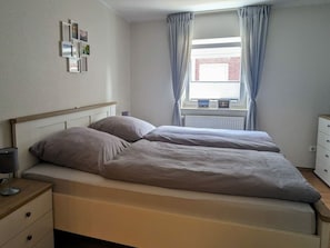 Ein Schlafzimmer mit einem Doppelbett, einem Nachttisch mit Lampe, einer Kommode und einem Fenster mit Vorhängen.