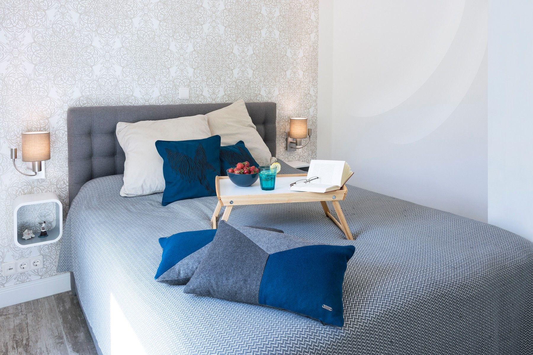 A neatly arranged bedroom with a bed, pillows, bedside lamps, a bedside table with a book and a glass of water, and a wall with a patterned wallpaper.