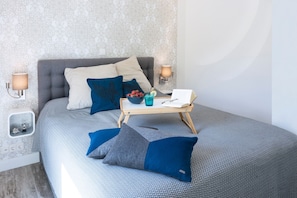 A neatly arranged bedroom with a bed, pillows, bedside lamps, a bedside table with a book and a glass of water, and a wall with a patterned wallpaper.