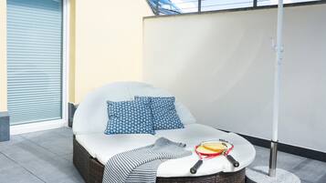 A sunlit outdoor seating area with a cushioned bench, a woven basket, and a tennis racket.