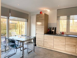 A modern kitchen with a glass dining table, metal chairs, wooden cabinets, and a window with a view of trees. - Zo10 Holiday Home Zoutelande (Zoutelande)