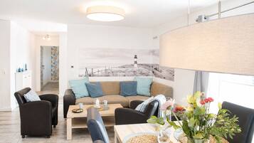 A modern living room with a sofa, chairs, a coffee table, and a dining table. There is a painting on the wall and a window with a view.