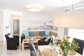 En moderne stue med sofa, stole, et sofabord og et spisebord. Et maleri hænger på væggen, og et vindue viser udsigt.