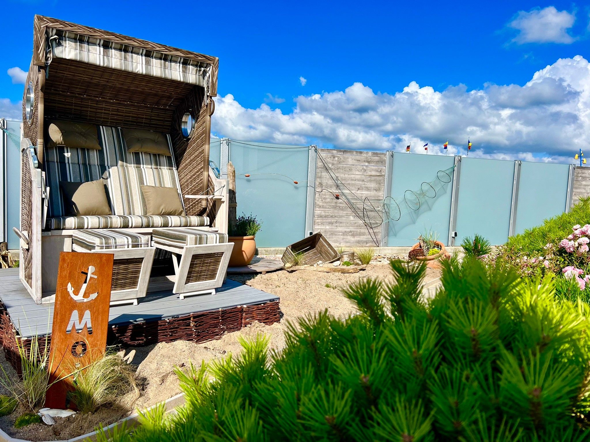 A beachside lounge with striped cushions and a wicker chair, a wooden deck, and a sign with an anchor symbol.