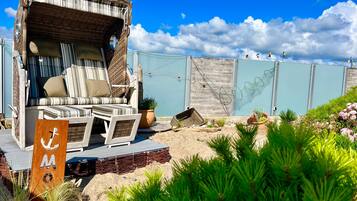 A beachside lounge with striped cushions and a wicker chair, a wooden deck, and a sign with an anchor symbol.