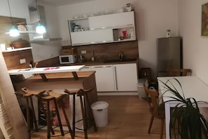 Cocina compacta con armarios blancos, microondas, lavamanos y comedor con sillas de madera.