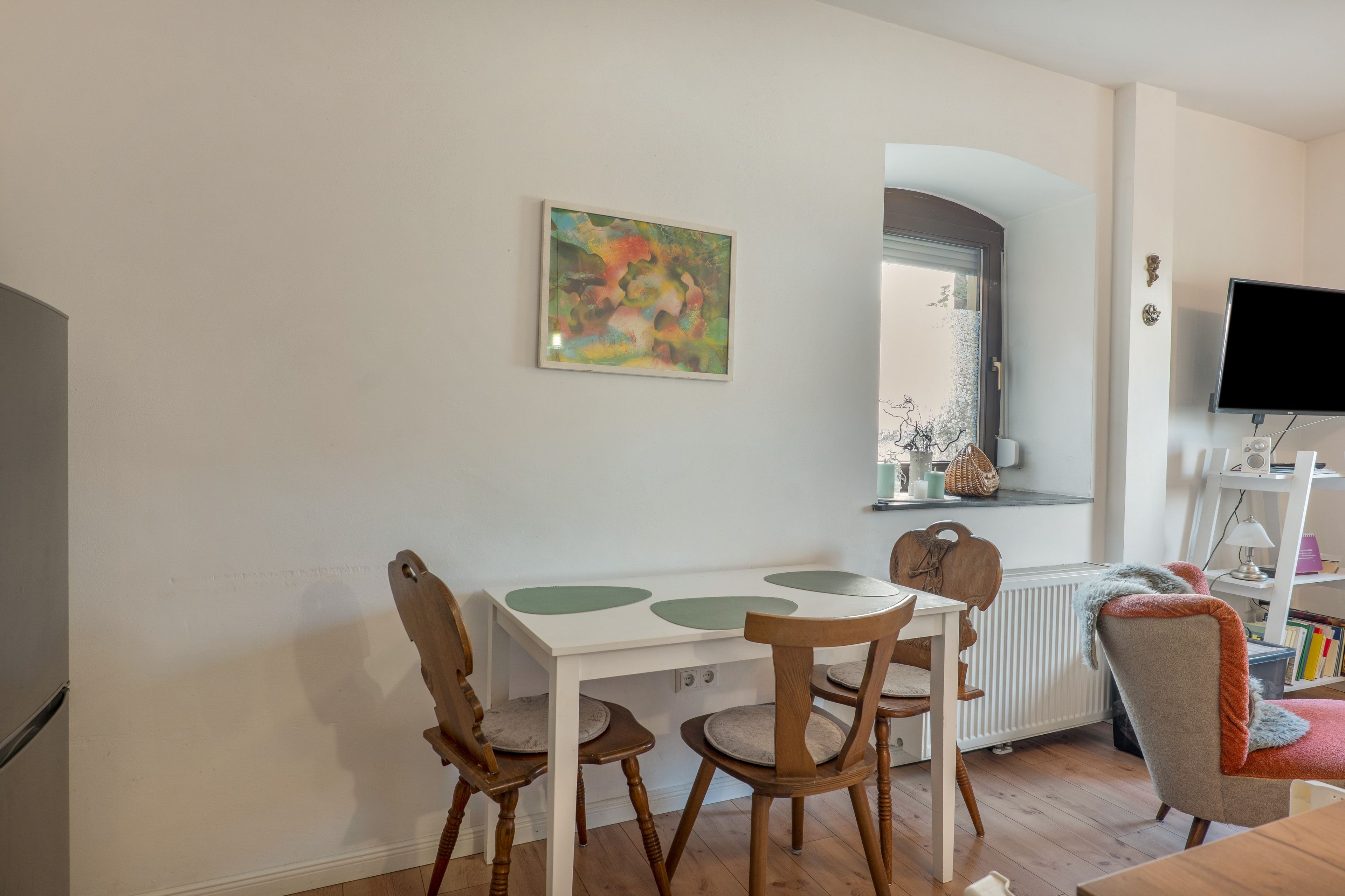 A dining area with a white table and chairs, a painting on the wall, and a television on a stand.