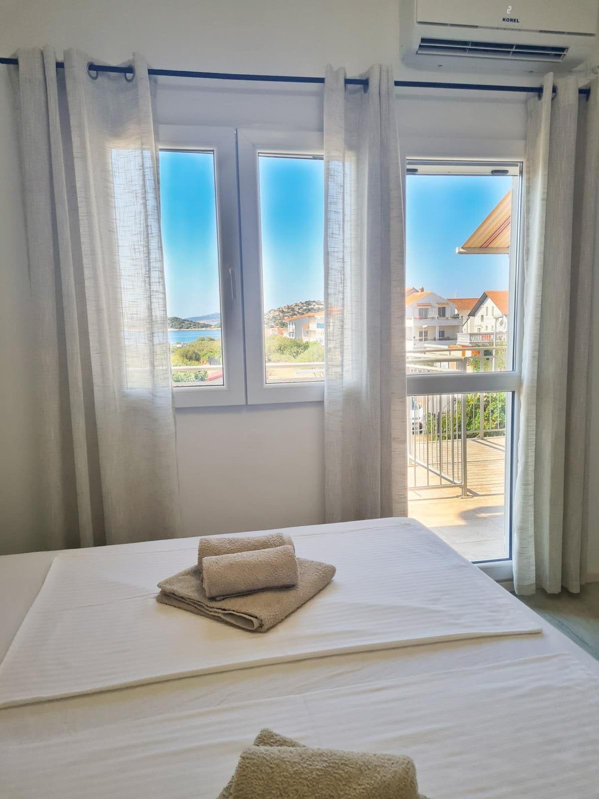 Een hotelkamer met een bed, uitzicht op zee en gebouwen, en een balkon met een hekwerk.