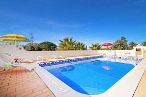 Une piscine entourée de palmiers, avec des chaises longues et des parasols, sous un ciel dégagé.