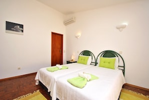 A hotel room with two beds, a wooden door, a wall-mounted air conditioner, and a small bedside table with a lamp.