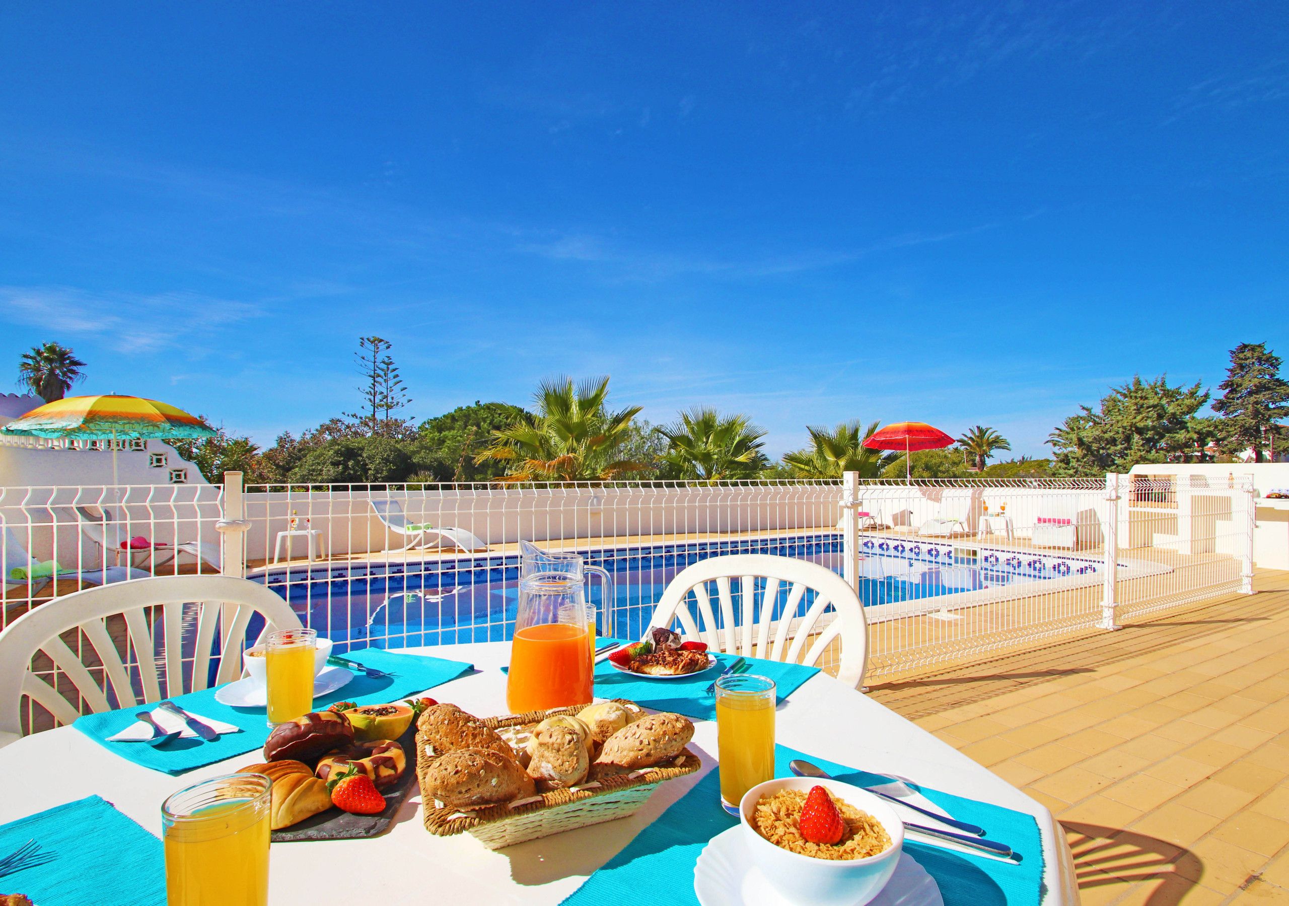 A poolside dining area with a table set for breakfast, including bread, fruit, and a beverage.