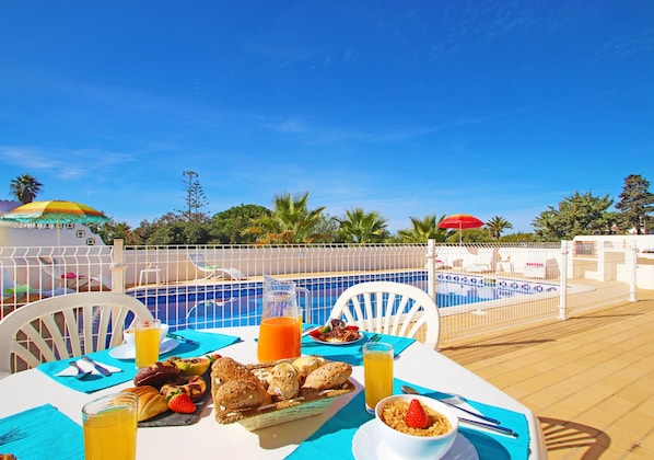 A poolside dining area with a table set for breakfast, including bread, fruit, and a beverage.
