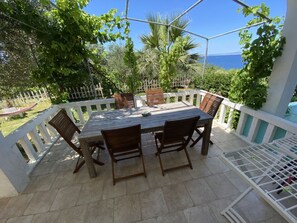An outdoor patio with a wooden table and chairs, surrounded by a white railing, with a view of the sea and greenery. - Villa Agata Ground Floor With Pool & Sauna (Rab)