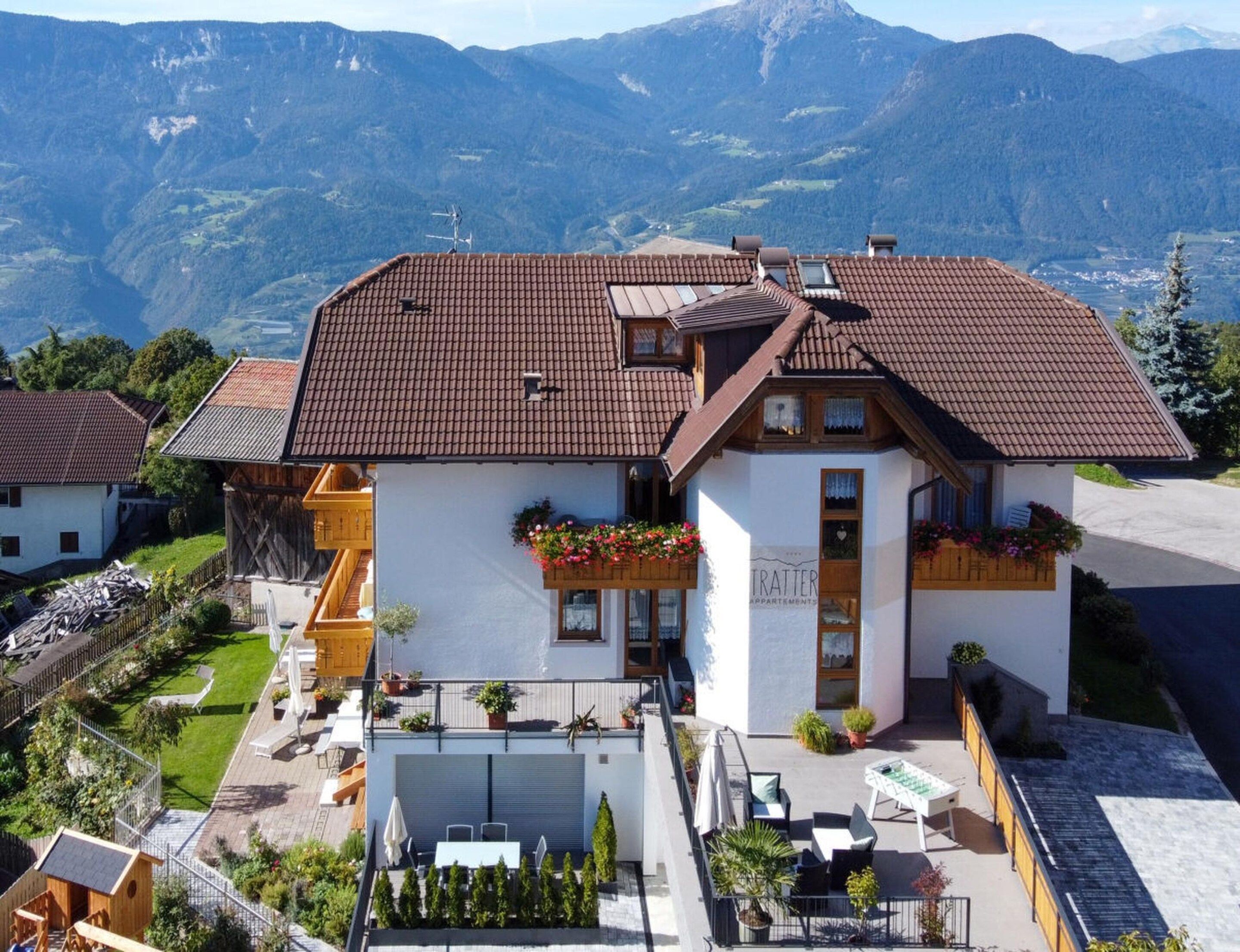 A two-story building with a brown tiled roof, a balcony with potted plants, and a view of mountains in the background.