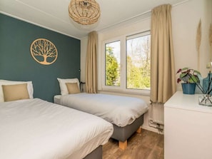 Ein Schlafzimmer mit zwei Betten, einem Fenster mit Vorhängen, einer Wanddekoration mit Baum-Motiv und einem Nachttisch mit Lampe und Topfpflanze.