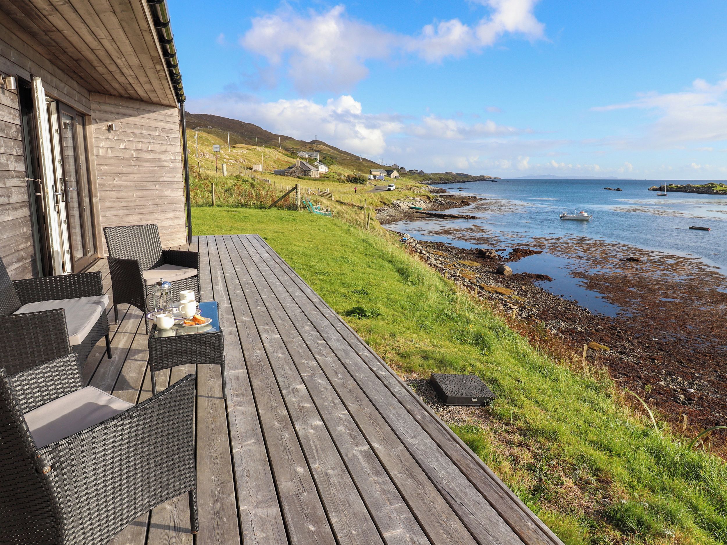 A wooden deck with outdoor furniture overlooking a coastal area with boats and a rocky shoreline.