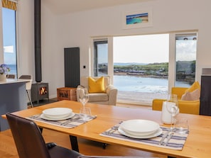 A dining area with a table set for two, a sofa with yellow cushions, a fireplace, and a view of the sea through large windows. - Holm (Harris)