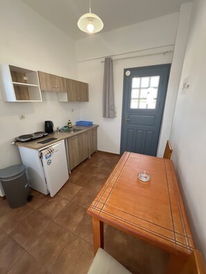 A compact kitchen with a small refrigerator, a coffee maker, and a sink. There is a wooden table with a chair, and a blue door with a window. - Studio Apartment 'Double Room - 202' with Sea View, Wi-Fi and Air Conditioning (Εμπορείο)