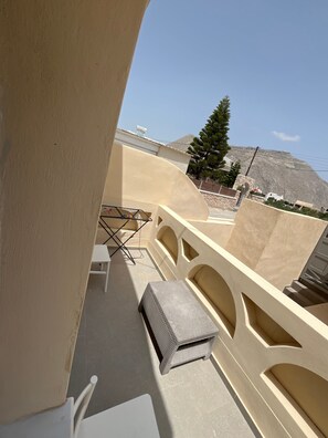 Ein Balkon mit einem weißen Stuhl, einem kleinen Tisch und einem Korbsitz. Im Hintergrund sind ein Baum und ein Berg zu sehen.