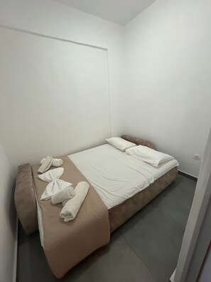 Ein Einzelbett mit beiger Matratze und weißen Leinentüchern, ein Nachttisch mit Handtuch sowie eine schlichte weiße Wand.
