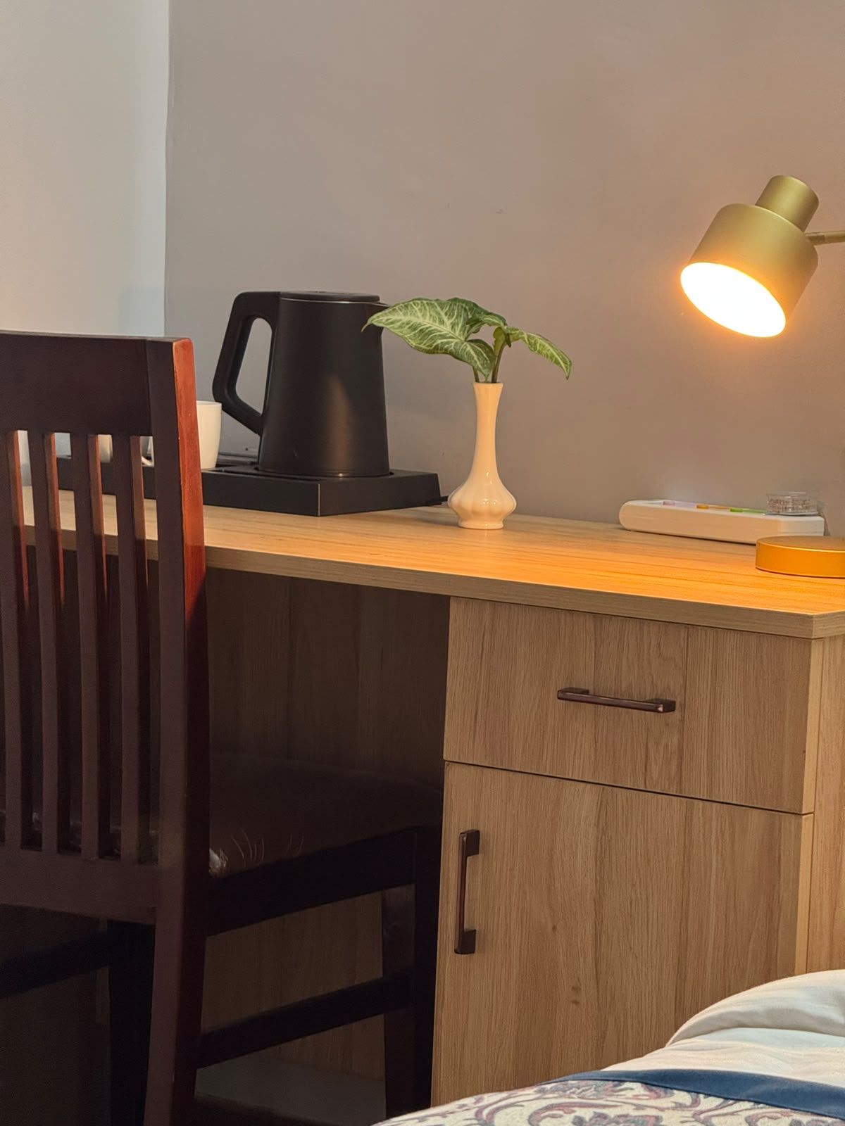 A wooden desk with a black coffee maker, a white vase with a plant, and a desk lamp.