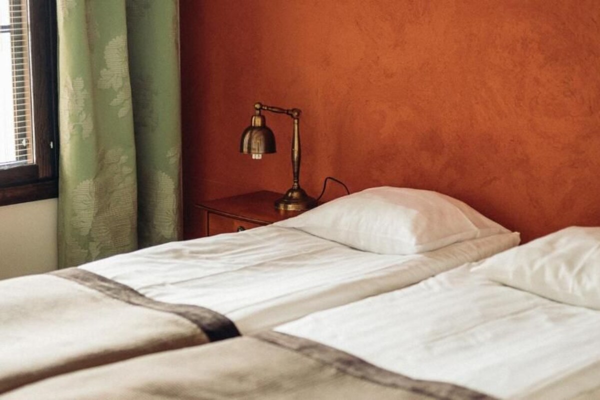 Ein Zimmer mit zwei Betten, einem Nachttisch mit Lampe und einem Fenster mit Vorhängen.