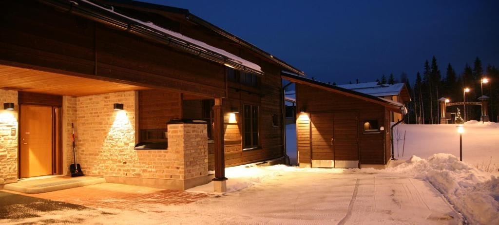 A wooden cabin with a stone foundation, a covered porch, and a snow-covered driveway.