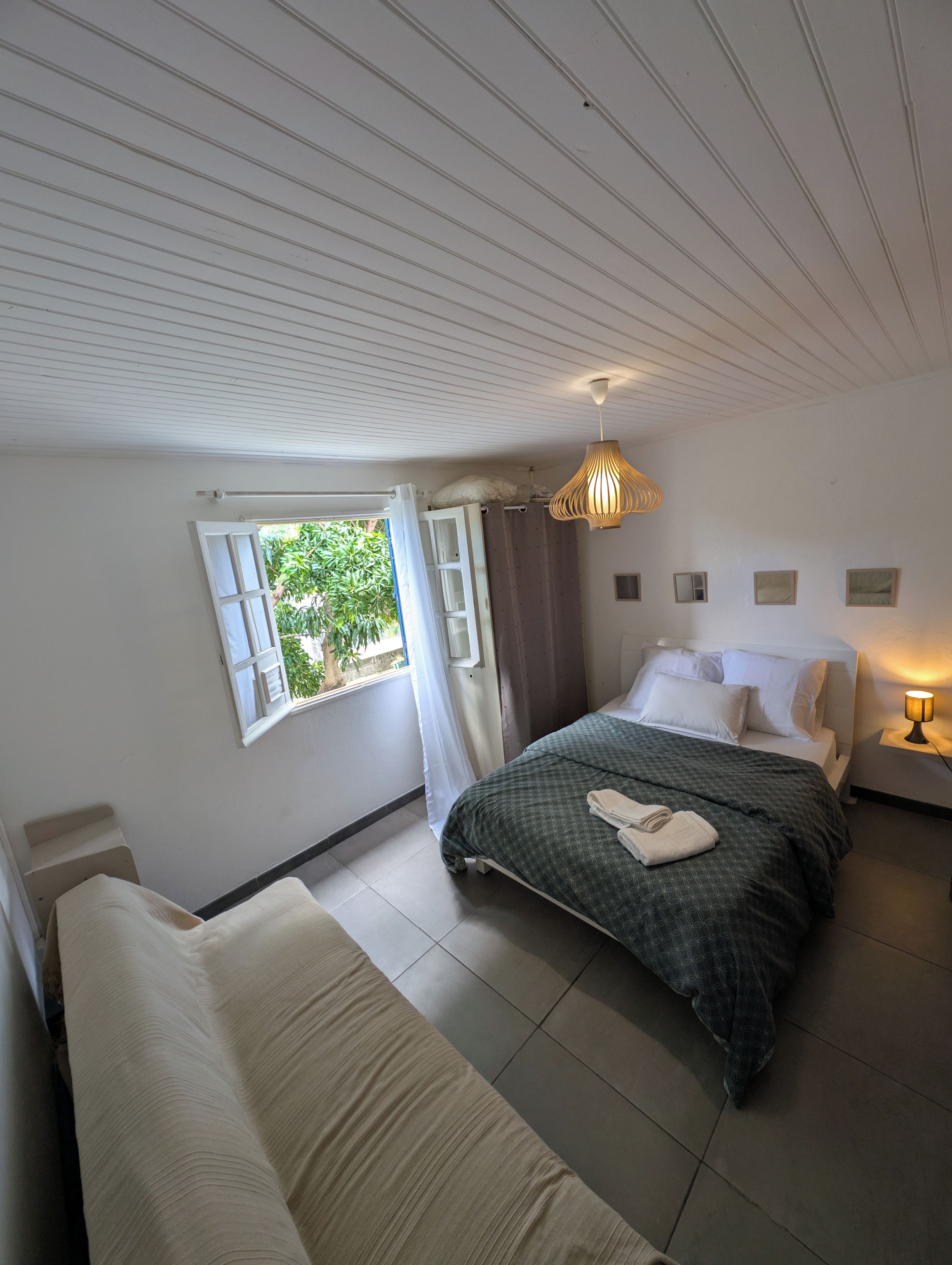A bedroom with a bed, a sofa, a window with curtains, a ceiling fan, and a bedside lamp.