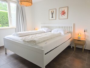 Ein weißes Bett mit Kopfteil, ein Nachttisch mit Lampe und zwei gerahmte Bilder an der Wand.