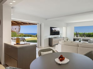 A modern living room with a sofa, a dining table with fruit, a TV, and a view of the ocean. - Holiday Home With 1 Bedroom (Arzachena)