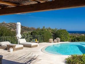 A poolside area with lounge chairs and a small table, overlooking a mountainous landscape and the sea. - Holiday Home With 1 Bedroom (Arzachena)