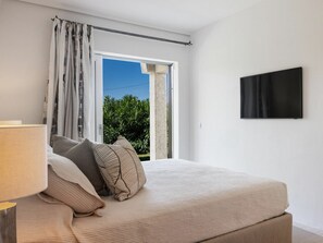 A bedroom with a bed, a window with curtains, a flat-screen TV on the wall, and a view of greenery outside. - Holiday Home With 1 Bedroom (Arzachena)
