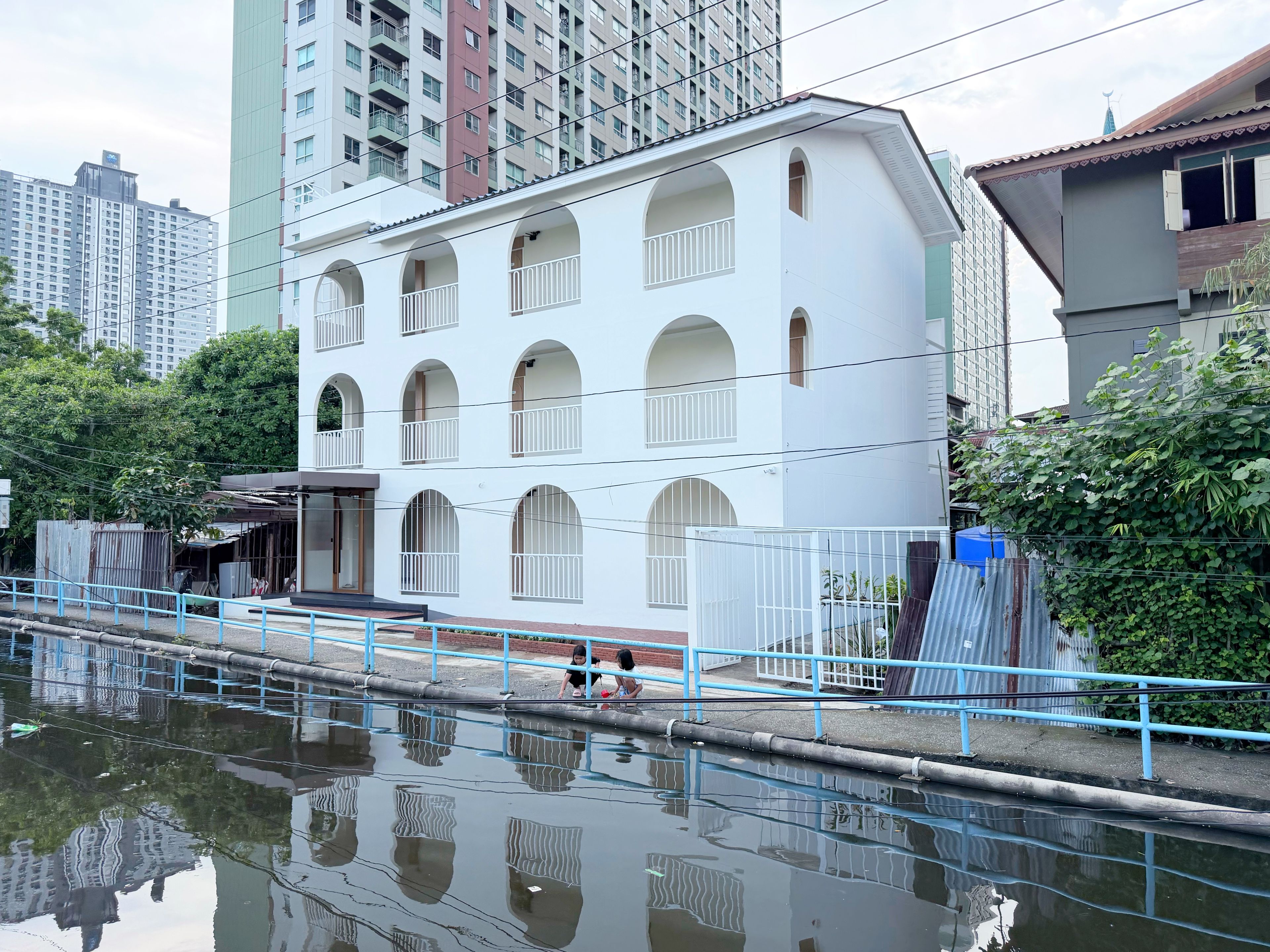 Un edificio de varios pisos con ventanas arqueadas y balcones, junto a una masa de agua con barandilla azul.