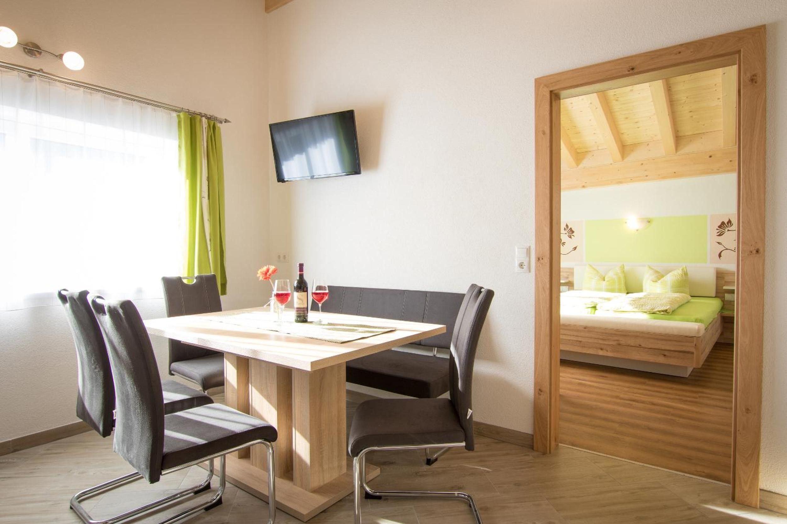 A room with a wooden dining table, four chairs, a television mounted on the wall, and a bed with a wooden headboard.