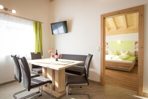Ein Zimmer mit einem Holztisch, vier Stühlen, einem an der Wand montierten Fernseher und einem Bett mit Holzvorhang.