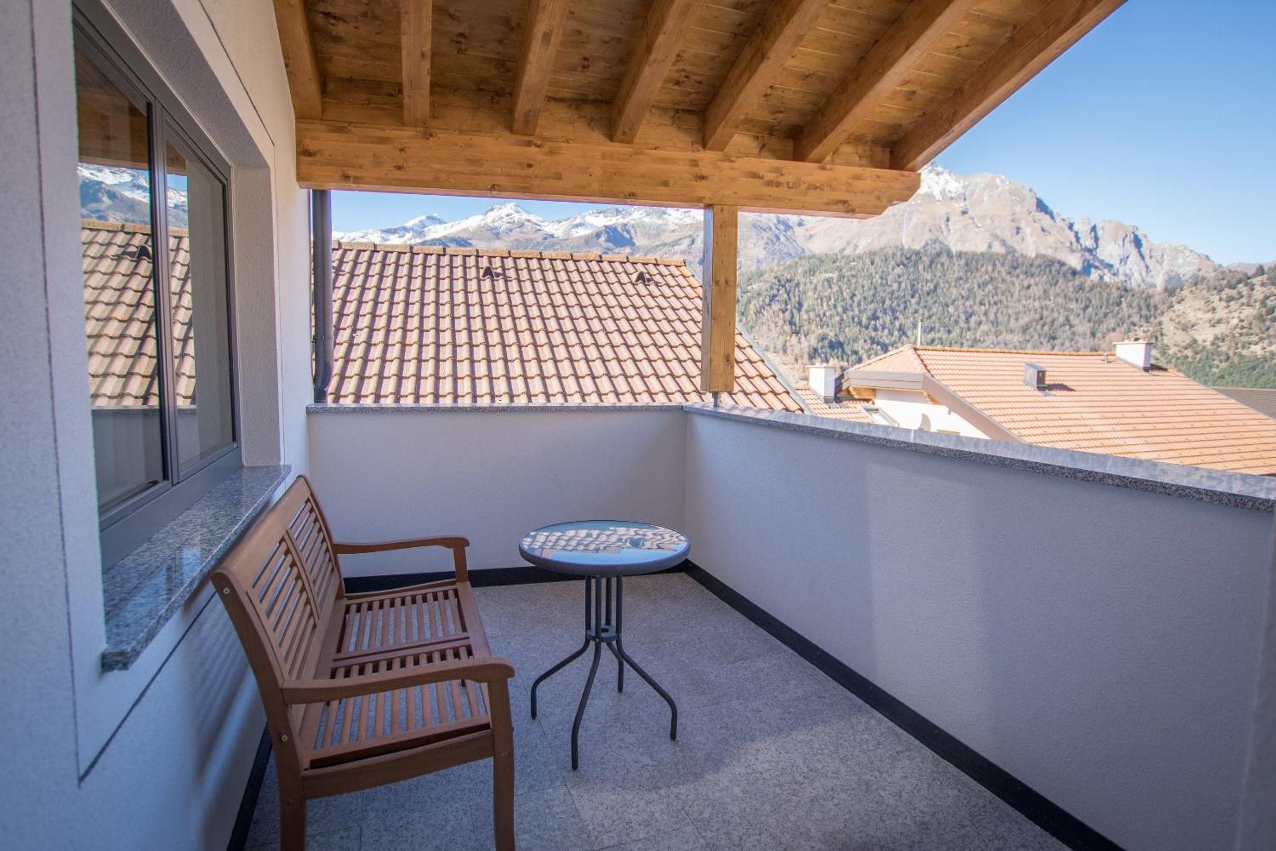Een balkon met een houten bankje en een tafeltje, met uitzicht op een bergachtig landschap met pannendaken.