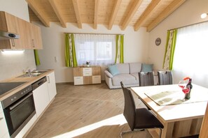 Una cocina moderna con armarios de madera y horno empotrado, un área de comedor con mesa y sillas, y una sala de estar con sofá y cortinas verdes.