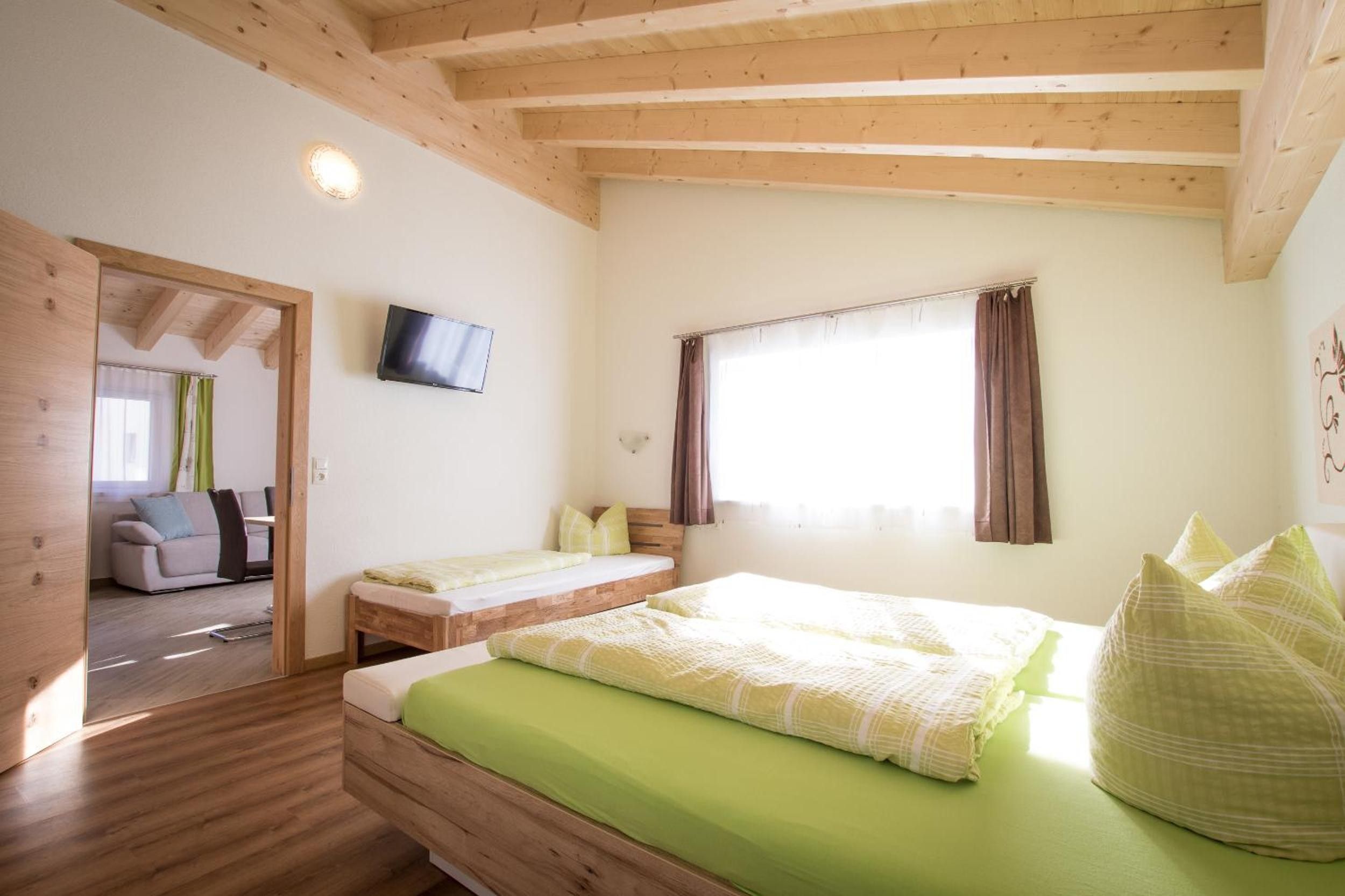 A bedroom with a wooden ceiling, a bed with green bedding, a television on the wall, and a window with curtains.