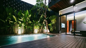 A swimming pool with a wooden deck, surrounded by lush tropical plants and a modern building with a glass entrance. - Moonstone Luxury Villa (Badung)